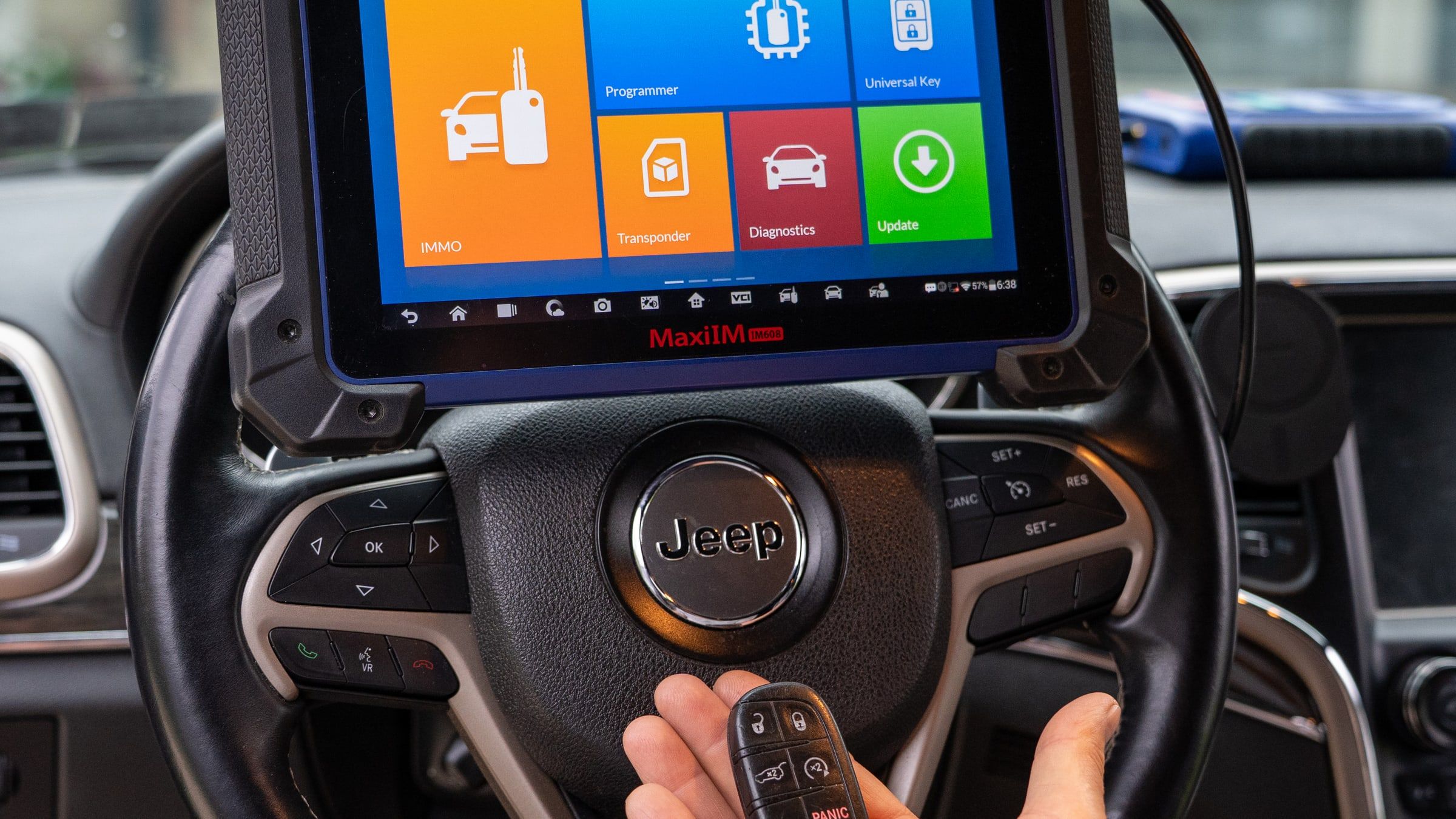 A key-programming tool mounted on a steering wheel showing its main menu, with a car key fob held below it.