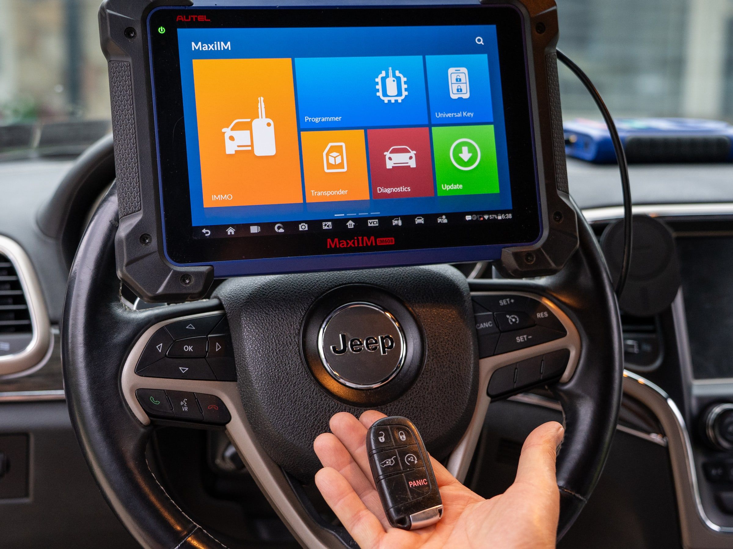 A key-programming tool mounted on a steering wheel showing its main menu, with a car key fob held below it.