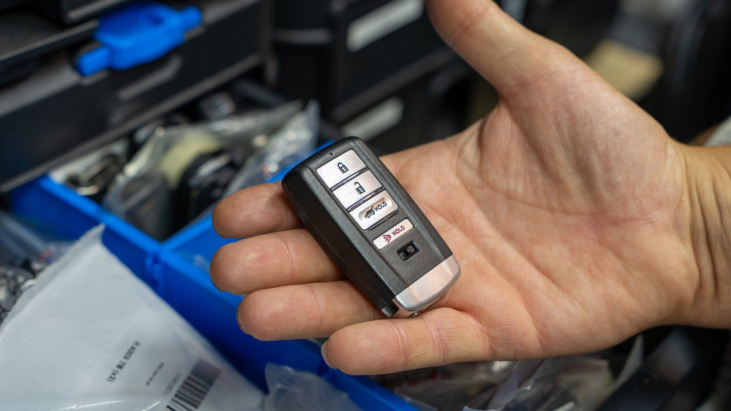 A smart key fob held in an open hand above a bin of packaged key blanks.